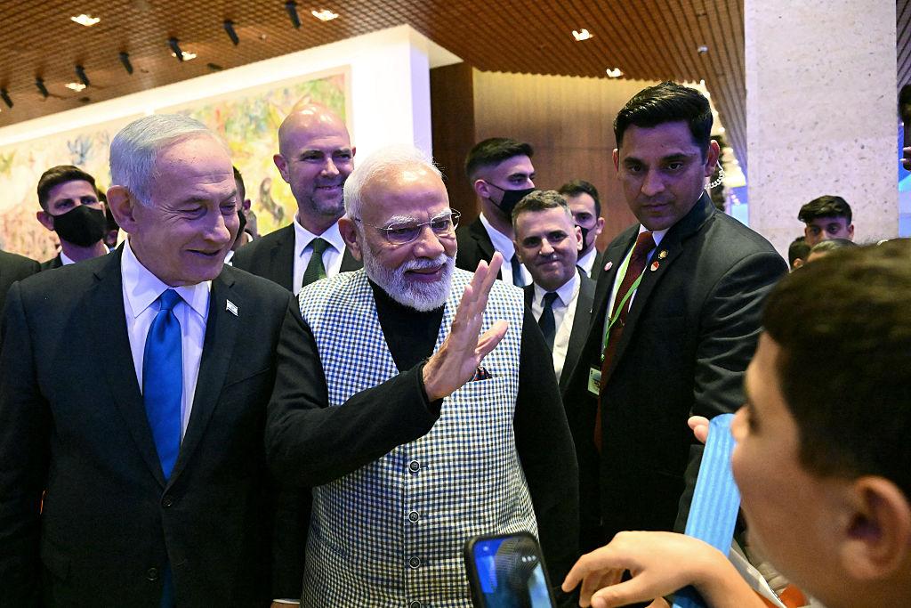 Indian Prime Minister Narendra Modi greets attendees alongside Israeli Prime Minister Benjamin Netanyahu at an indoor event, surrounded by officials and security personnel, as a person in the foreground holds up a smartphone to take a photo.
